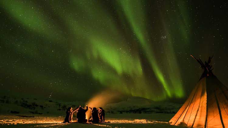 A group of people by the bonfire outside the Lavvo, underneath the northern lights. Photo.